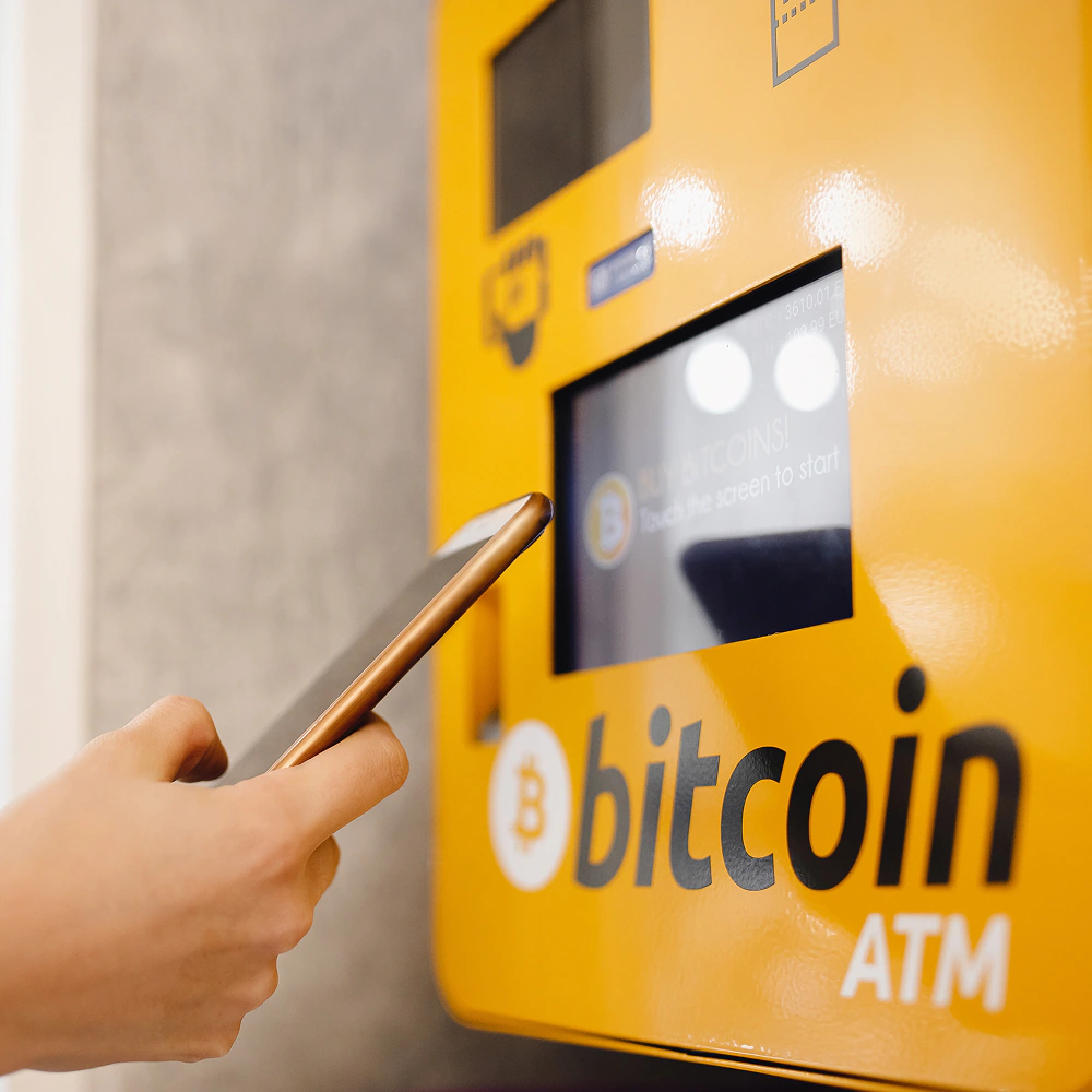 Osoba korzysta z bitomatu (bitcoin ATM) i trzyma smartfon przy ekranie urządzenia